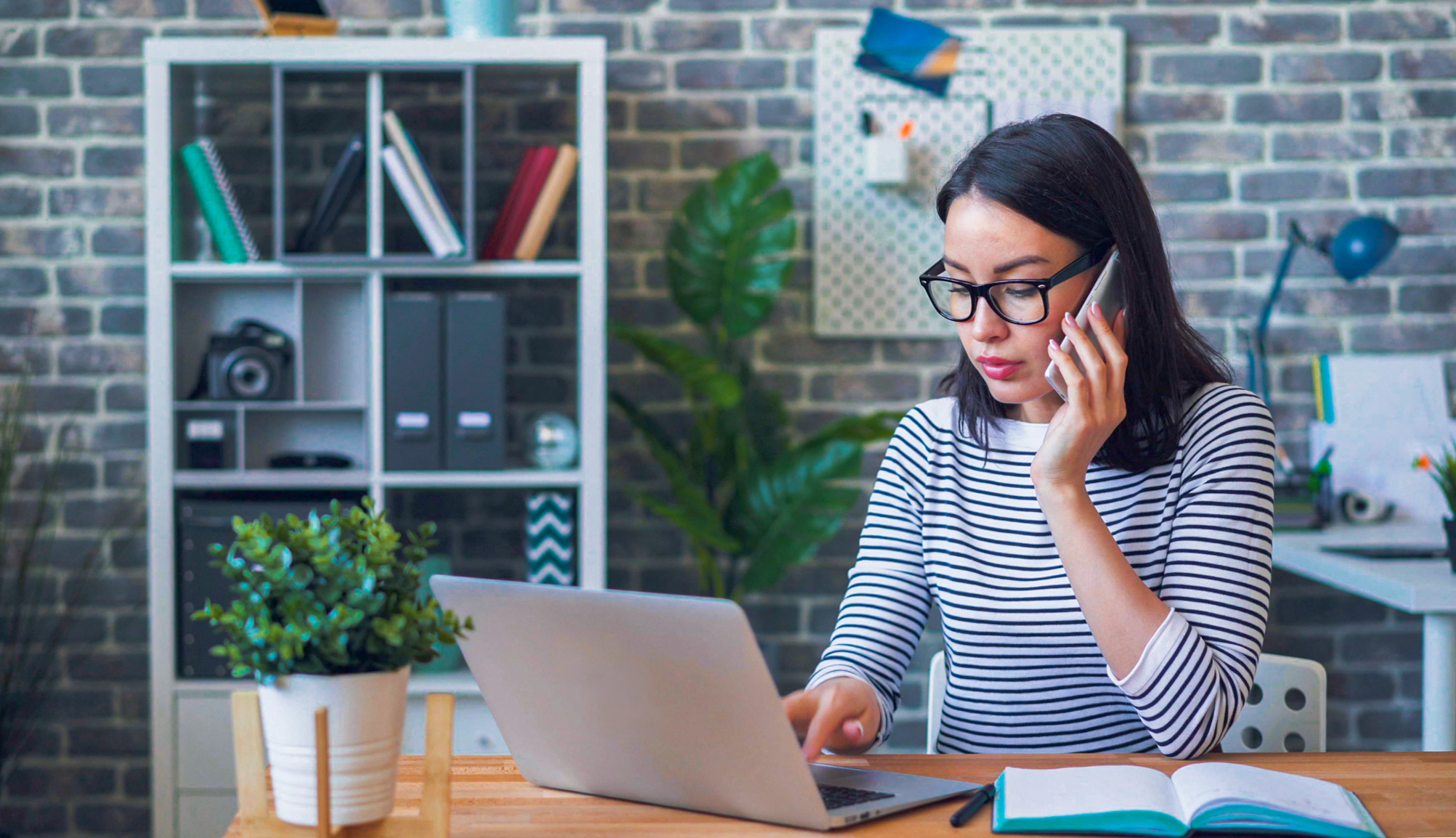 Mulher trabalhando remotamente, usando notebook e celular para contratar seguro empresarial, com agenda e materiais de escritório sobre a mesa