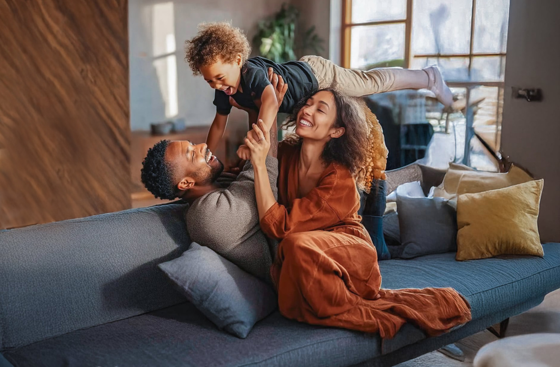 Família sorridente brincando com o filho no sofá de casa iluminada pelo sol
